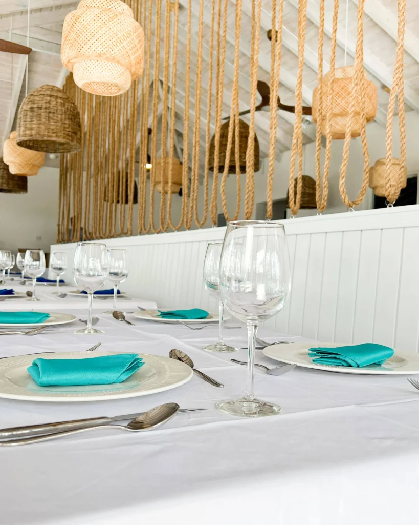 Close-up of a table setting with a white tablecloth, turquoise napkins on plates, and wine glasses, with hanging rope pendant lights in the background.