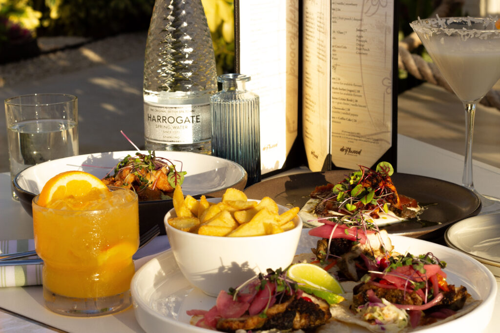Table spread featuring a vibrant orange drink with citrus slices, a bowl of fries, and a plate of appetizers topped with pickled onions and microgreens, with a martini glass, menu, and water bottle in the background.