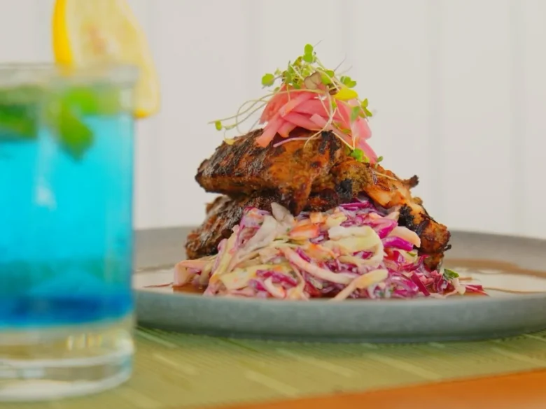 Plate with a stacked grilled entrée atop a bed of colorful coleslaw salad, served on a green placemat with a beverage in the background.