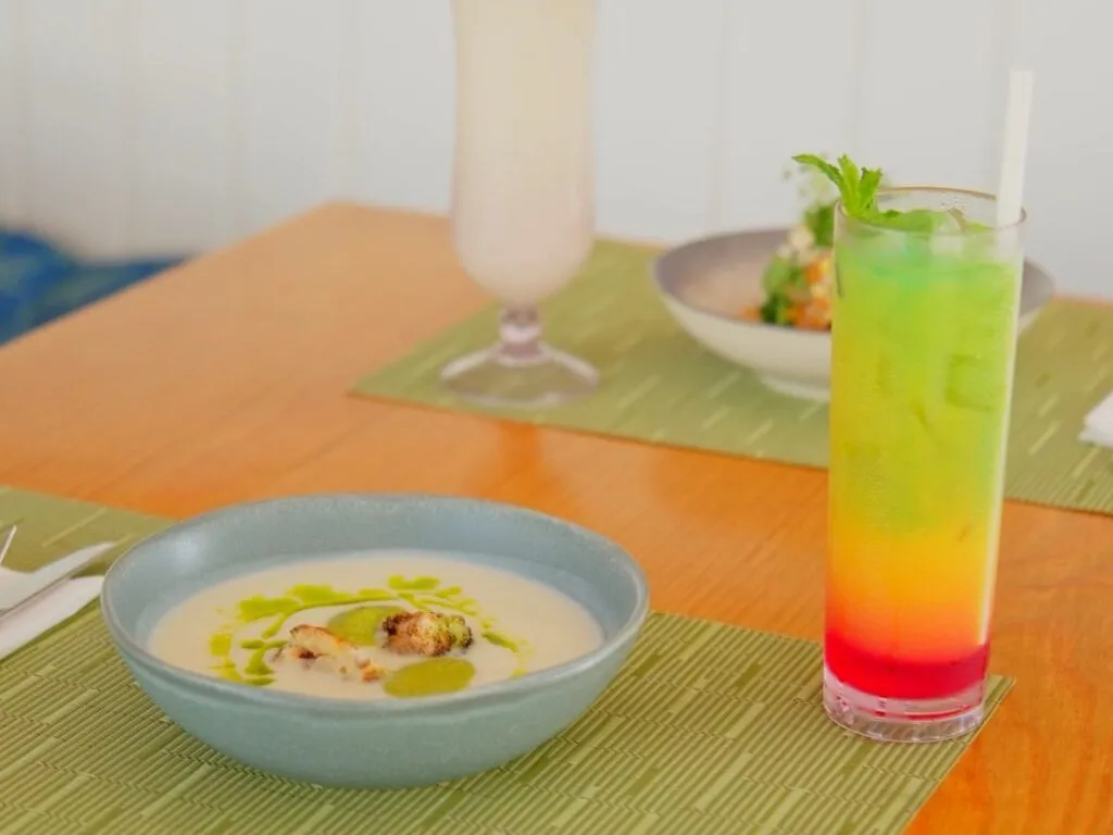 Colorful layered cocktail and a bowl of creamy soup with small pieces, set on a table with other drinks and a salad in the background.