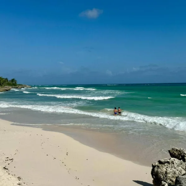 Sandy beach with turquoise waves and rocks, with two people wading in the water and a house and trees in the distance.