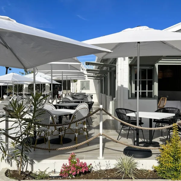 Outdoor dining area featuring multiple tables and chairs under large white umbrellas on a paved patio, with surrounding plants and the hotel building in the background.