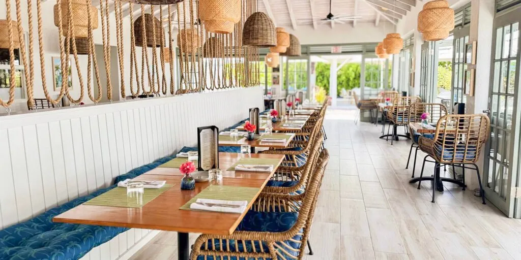 Interior dining area at Driftwood Restaurant featuring a long bench with wooden tables and wicker chairs, decorated with rope pendant lights and large windows letting in natural light.