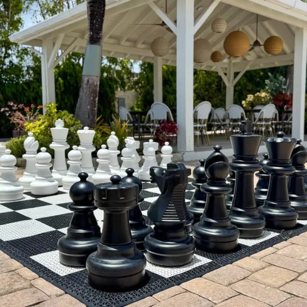 Outdoor area featuring a large chess board with oversized black and white chess pieces on a paved patio, with a gazebo and seating area in the background.