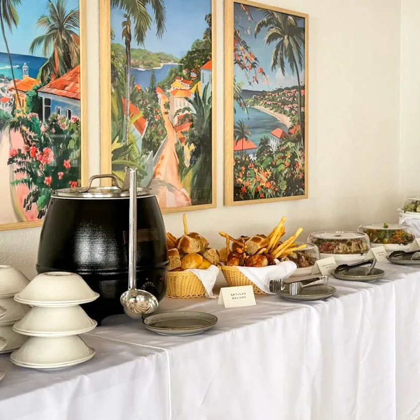 Buffet table with a black soup kettle, bowls, bread and pastries, and other dishes, with colorful paintings of coastal scenes on the wall.
