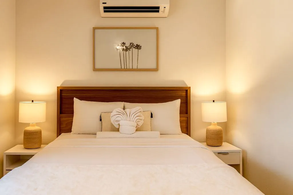Bright, cozy bedroom with neutral decor, featuring a wooden headboard, soft white bedding, a towel folded into a decorative shape, bedside lamps, and framed palm tree artwork above the bed.
