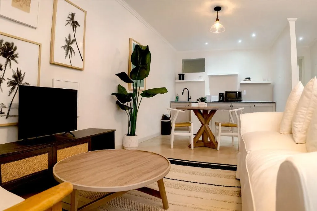 Open-plan living and kitchen area in the Garden View – Two Bedrooms suite at South Point Hotel, featuring a cream sofa with textured pillows, round wooden coffee table, large potted plant, framed palm artwork, flat-screen TV on a wood console, dining table with black chairs, and modern kitchenette with white cabinets and black fixtures.