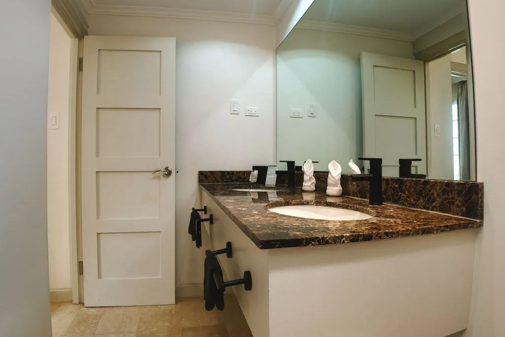 Modern bathroom with white double doors, large wall mirror, marble countertop with single sink, black fixtures, rolled white towels, and soft ambient lighting.