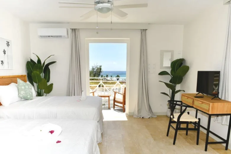 The bedroom of the Ocean View Twin Bed Suite. The suite's beds are made, with tropical decor throughout the room. A TV rests on a small desk, complete with bamboo chair, and the balcony overlooks the ocean and hotel grounds.