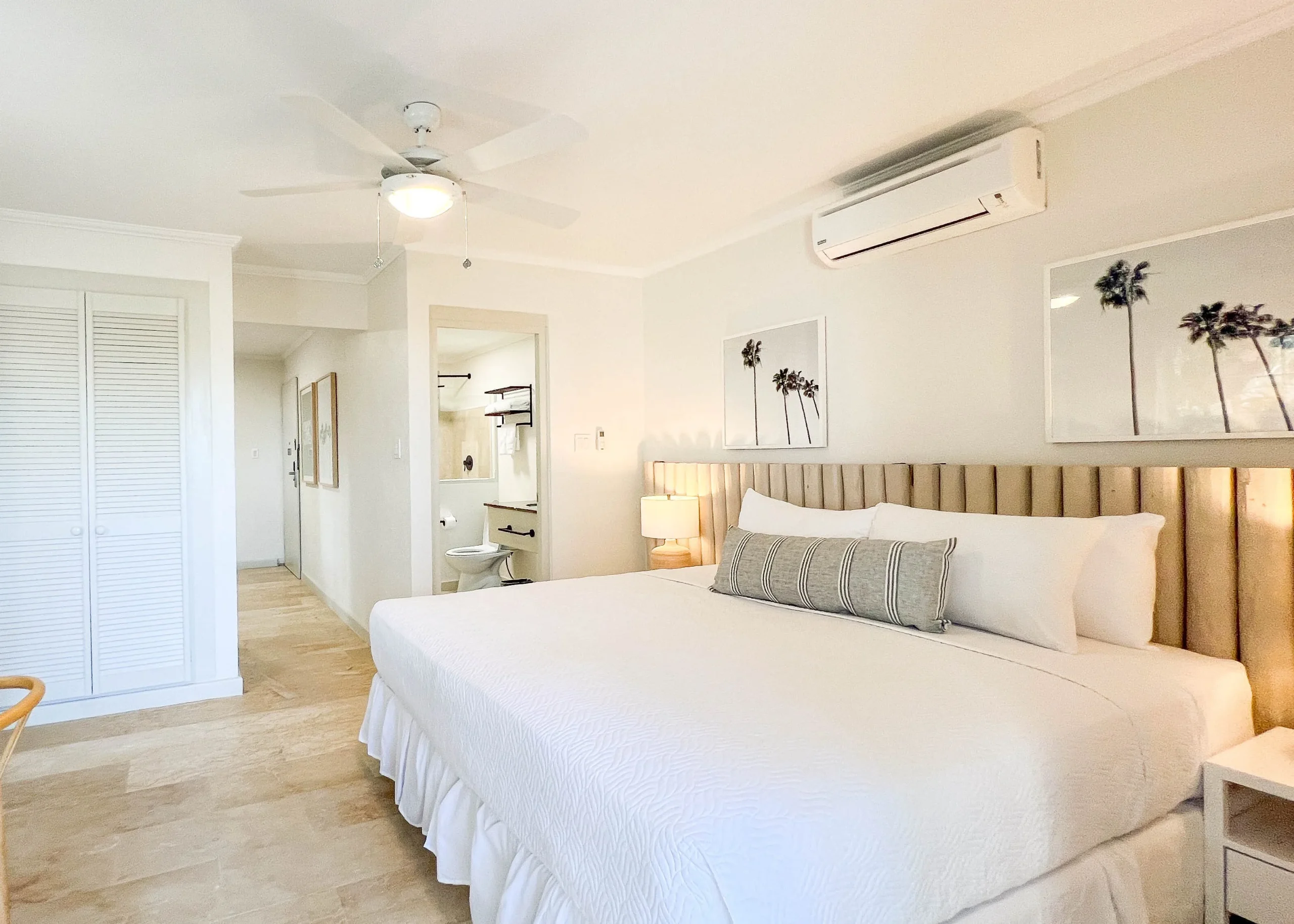 Spacious king bedroom in the Garden View King suite at South Point Hotel Barbados, featuring a plush king bed with white linens and textured bolster pillow, padded headboard wall, bedside lamps, ceiling fan, modern artwork, and bright natural light from a hallway leading to the bathroom and closet.