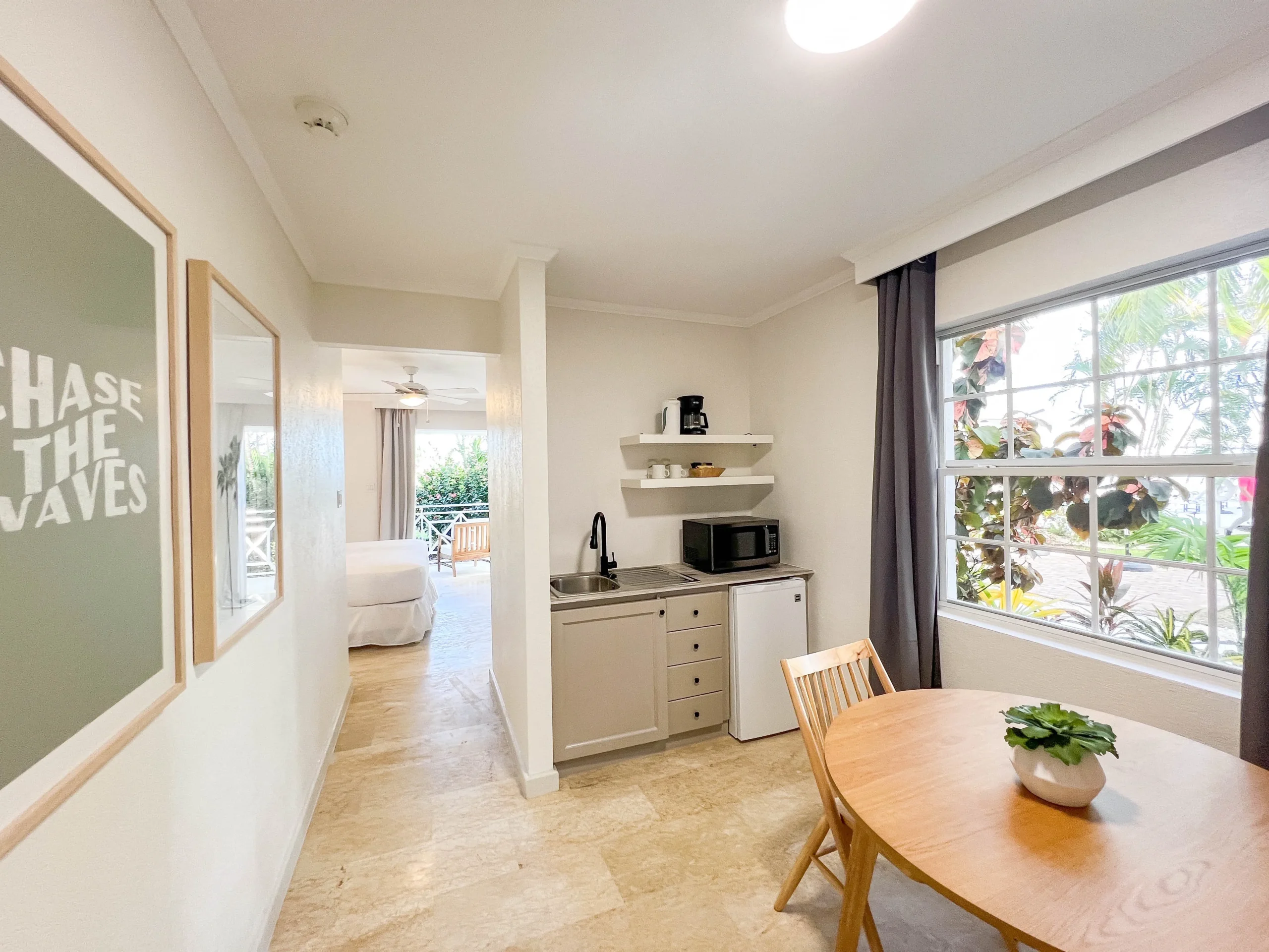 Bright kitchenette and dining area in the Garden View King suite at South Point Hotel Barbados, featuring white cabinetry with built-in microwave and sink, round wooden dining table with potted plant, large window with garden foliage outside, and hallway leading to the bedroom and patio.