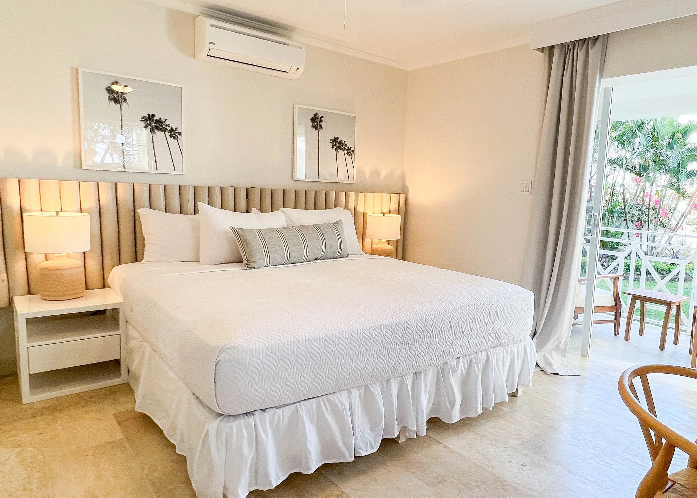 Bright king bedroom in the Garden View King suite at South Point Hotel Barbados, featuring a plush king bed with white linens and striped bolster pillow, bedside lamps, framed palm artwork, soft neutral decor, and sliding doors leading to a private balcony with tropical greenery.