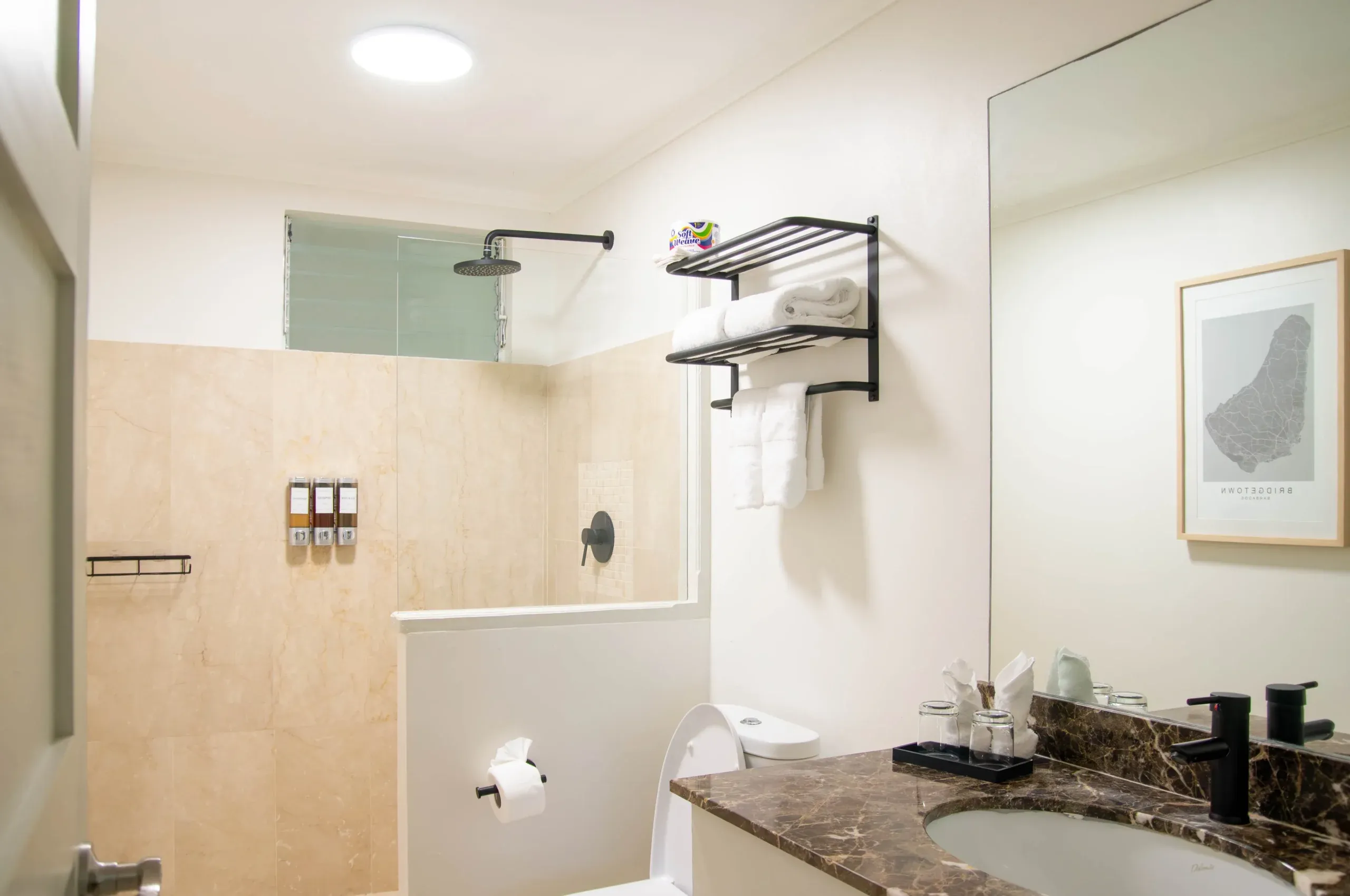 Modern bathroom in the Garden View Two Twin suite at South Point Hotel Barbados, featuring a walk-in glass shower with rain showerhead, wall-mounted bath amenities, towel shelf with folded white towels, and a marble vanity with sink and toiletries.
