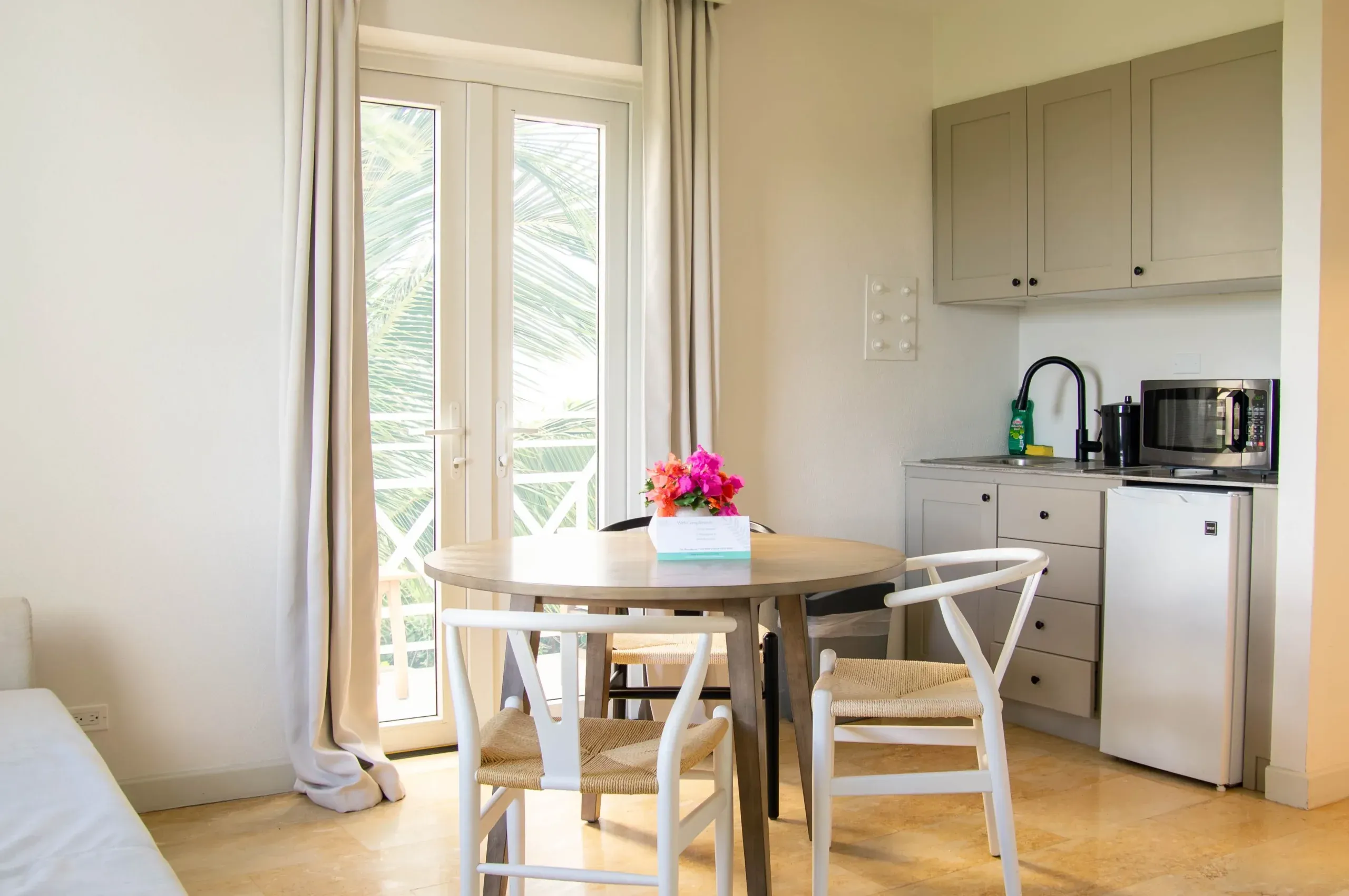 The kitchenette of the King Ocean View Suite, with a small table surrounded by chairs and topped by colourful flowers. Outside the closed glass door is a balcony overlooking the ocean.