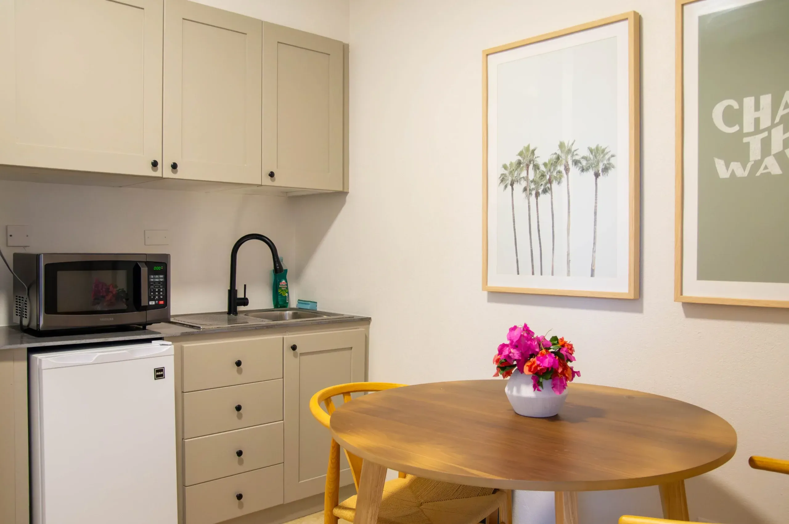 Kitchenette in a suite featuring beige cabinets, a black faucet, microwave, mini fridge, and a round wooden table with a vase of pink flowers, with framed palm tree artwork on the wall.