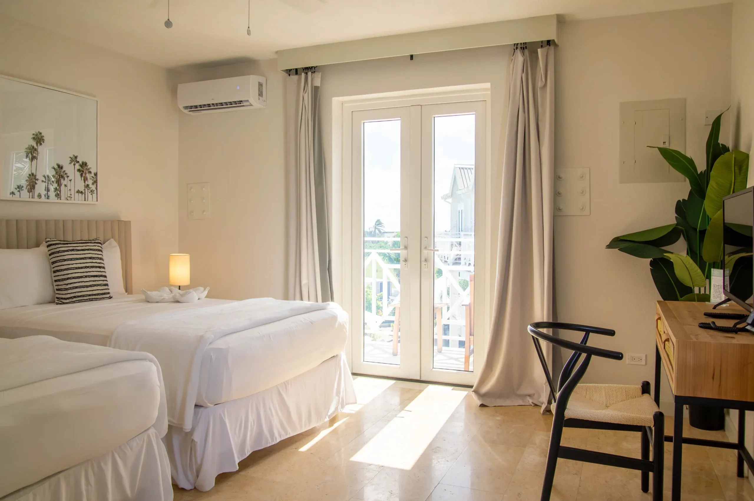Suite view of the Two Double Pool View. Two nicely-made beds and a desk are visible. The balcony leads to the pool of South Point Hotel Barbados.