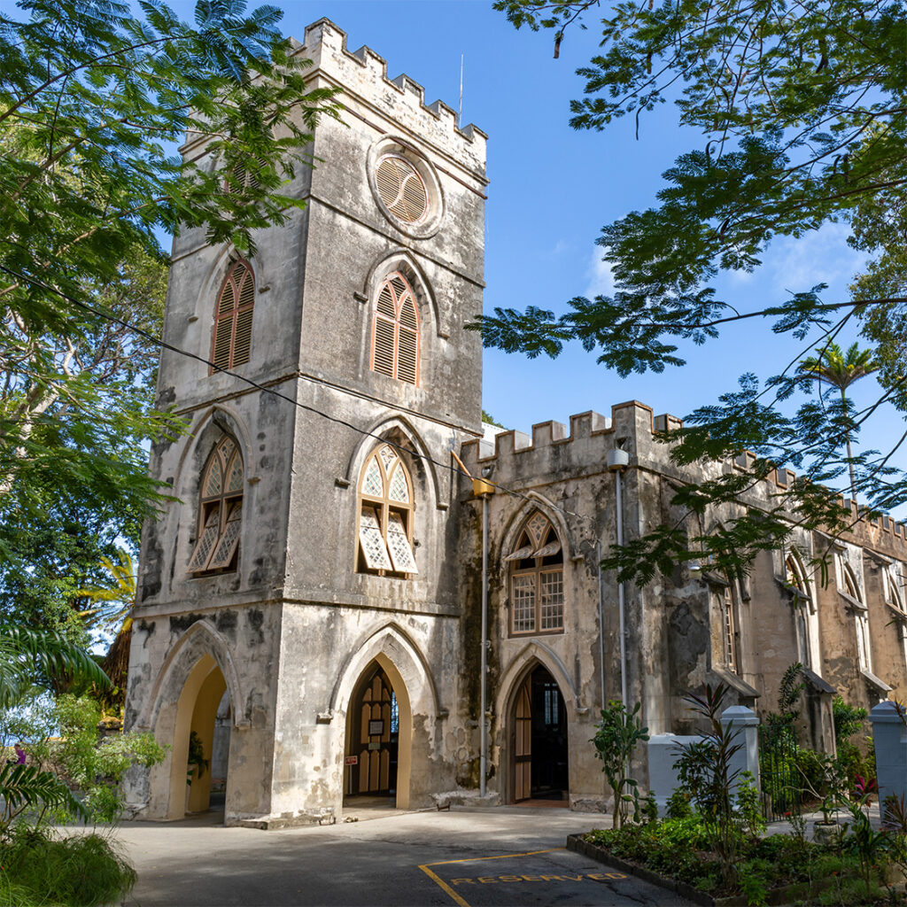 Stone Gothic-style church with arched windows and bell tower surrounded by trees in Barbados under a bright blue sky