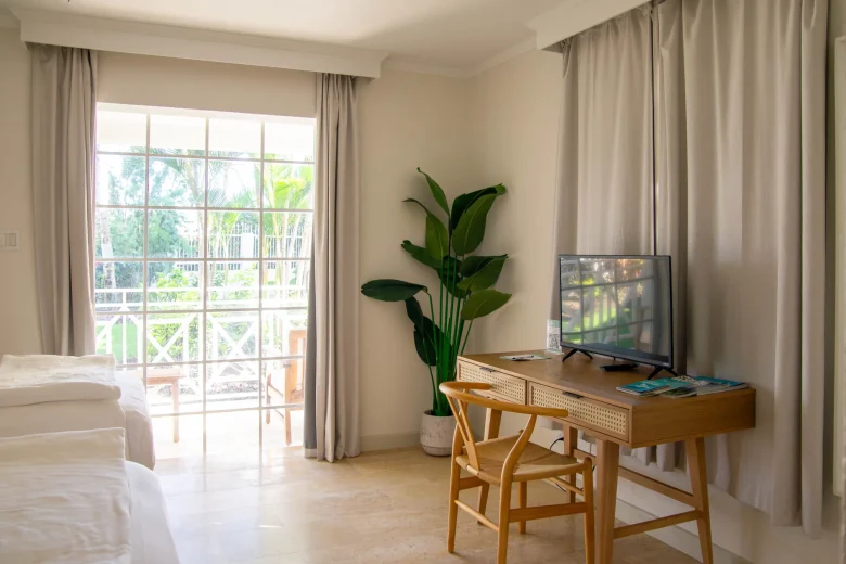 Bright twin bedroom in the Garden View Two Twin suite at South Point Hotel Barbados, featuring two white twin beds, a wooden writing desk with a flat-screen TV, a potted banana plant, large curtains framing garden-view windows, and a private furnished patio outside.