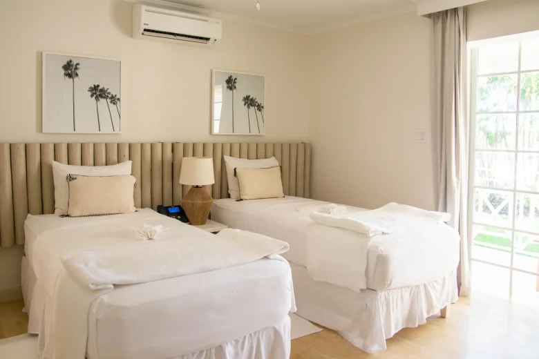 Bright twin bedroom in the Garden View Two Twin suite at South Point Hotel Barbados, featuring two neatly made single beds with white linens, tufted beige headboard wall, bedside tables with lamps, framed palm artwork, and a floor-to-ceiling window overlooking tropical gardens.
