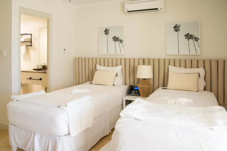 Bright twin bedroom in the Garden View suite at South Point Hotel Barbados, featuring two neatly made single beds with white linens, a padded headboard wall, bedside lamps, framed palm tree artwork, and a doorway leading to the bathroom.
