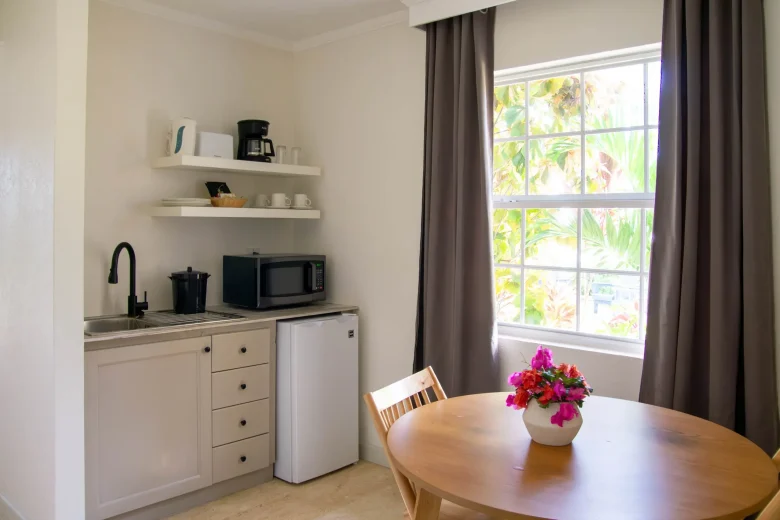 Modern kitchenette with white cabinets, stainless mini fridge, black microwave, coffee maker and kettle on wall shelves, round wooden dining table with bright pink flowers, and a large window overlooking palm trees.