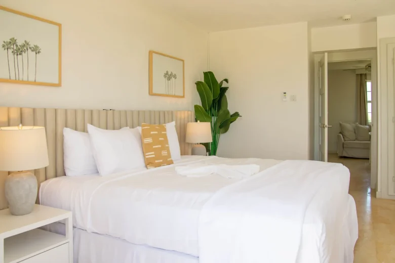 The king sized bed of the King Ocean View Suite, nicely made, and decorated with yellow & cream coloured decor. The sitting room is visible in the background.