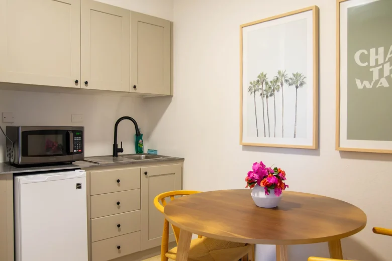 Kitchenette in a suite featuring beige cabinets, a black faucet, microwave, mini fridge, and a round wooden table with a vase of pink flowers, with framed palm tree artwork on the wall.