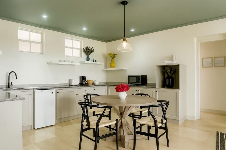 Kitchenette in a presidential suite featuring beige cabinets, a black faucet, microwave, mini fridge, and a round wooden table with a vase of red flowers, with pendant lights overhead and palm artwork on the wall.