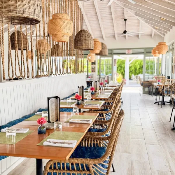 Interior dining area at Driftwood Restaurant featuring a long bench with wooden tables and wicker chairs, decorated with rope pendant lights and large windows letting in natural light.