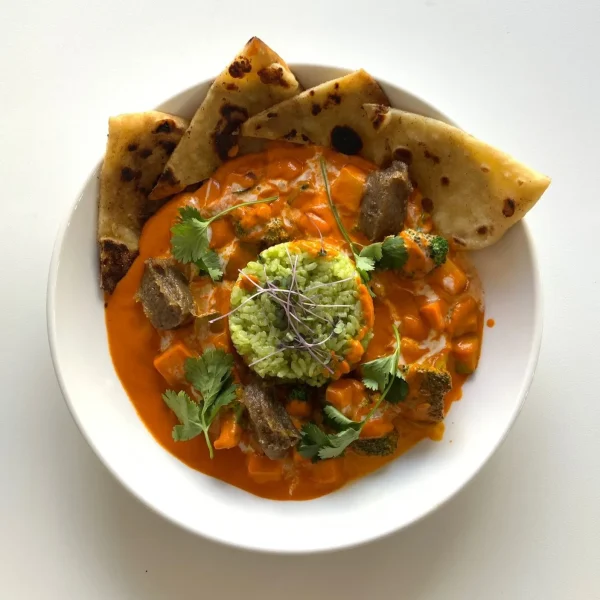 Bowl of savory orange dish topped with a green herb mixture, garnished with microgreens and cilantro, served with pieces of flatbread on the side.