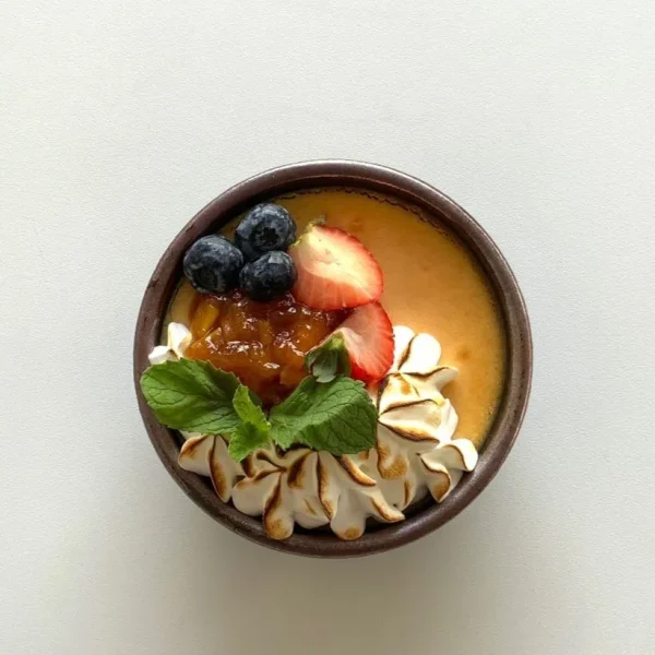 Dessert bowl featuring a creamy base topped with caramelized sauce, blueberries, strawberry slices, mint leaves, and piped meringue.