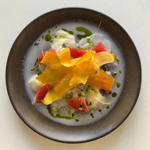 Plated dish featuring a creamy base with pieces of seafood or vegetables, topped with thin crispy chips and garnished with grapefruit segments and green drizzle.