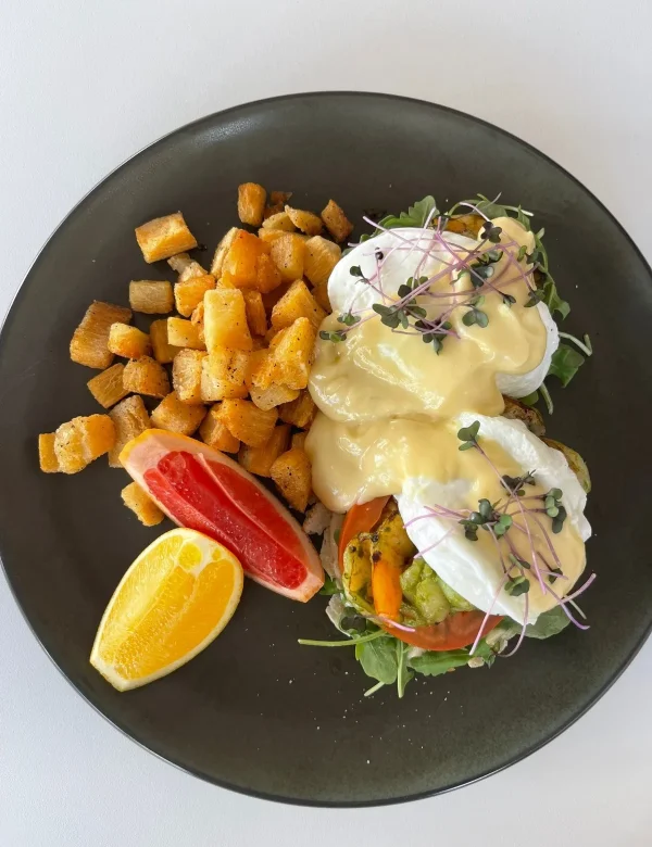 Plated brunch dish featuring two poached eggs with creamy sauce on toast with avocado and tomato, accompanied by roasted potato cubes and citrus wedges.